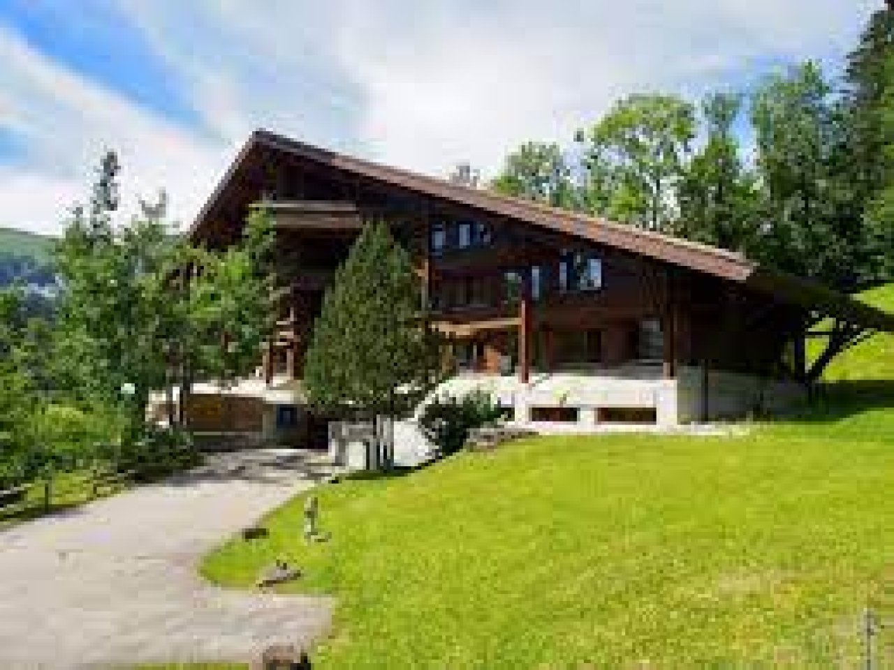 Ein dunkelbraun lasiertes Chalet mit einer kleinen Einfahrt, grünen Bäumen im Hintergrund und blauem Himmel.